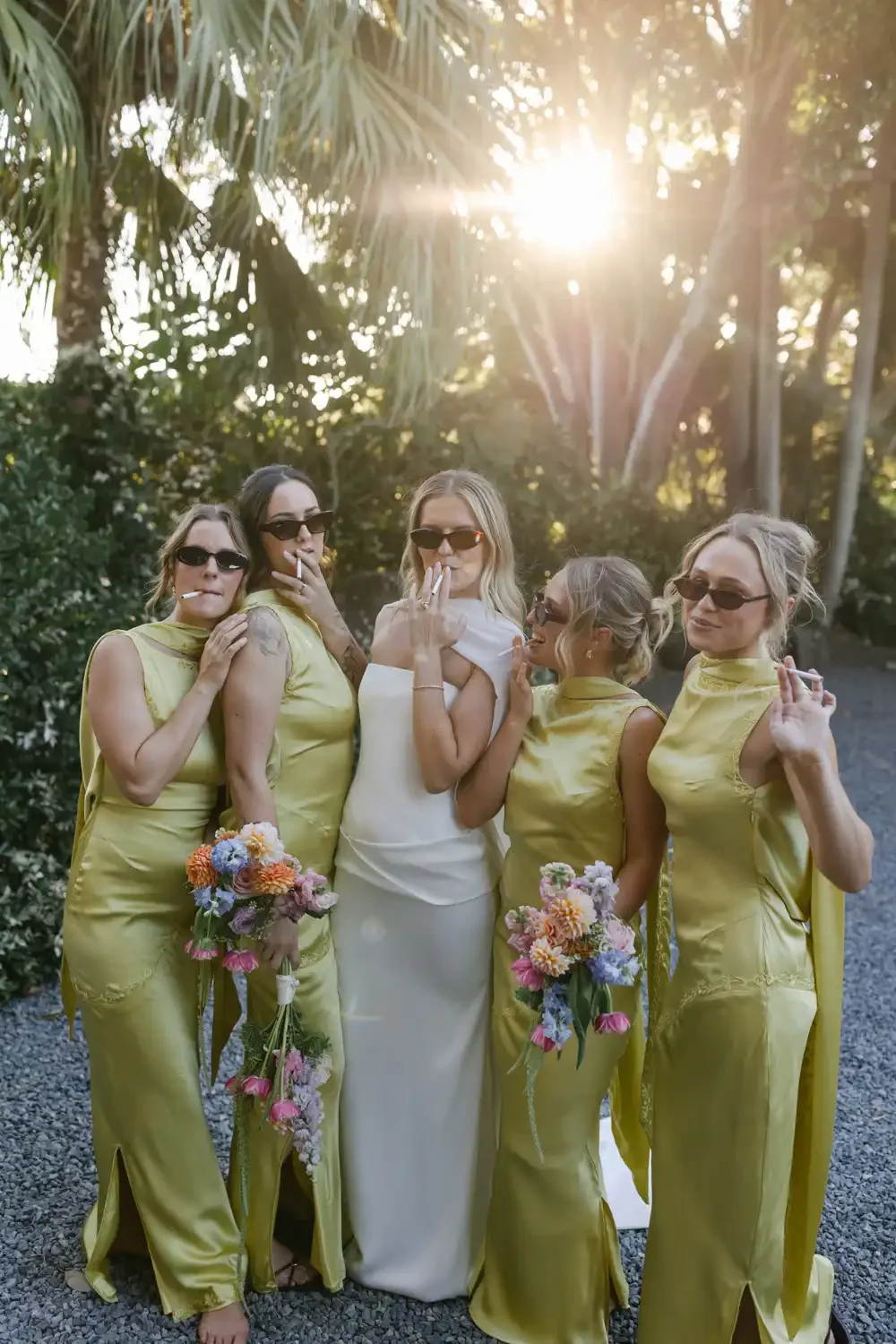 Five women, one in a white wedding dress and four in matching yellow satin dresses, standing outdoors in sunlight, holding bouquets of pink and purple flowers, wearing sunglasses, and smoking cigarettes during a wedding celebration.