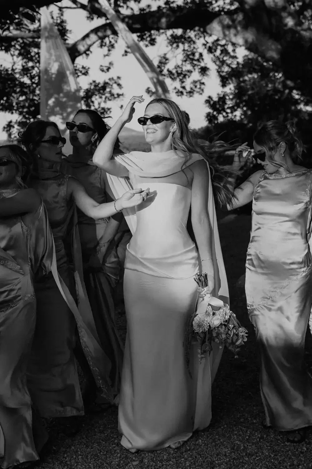 A group of women in elegant dresses and sunglasses, with one woman in a white gown holding a bouquet, smiling and waving, outdoors under a large tree.