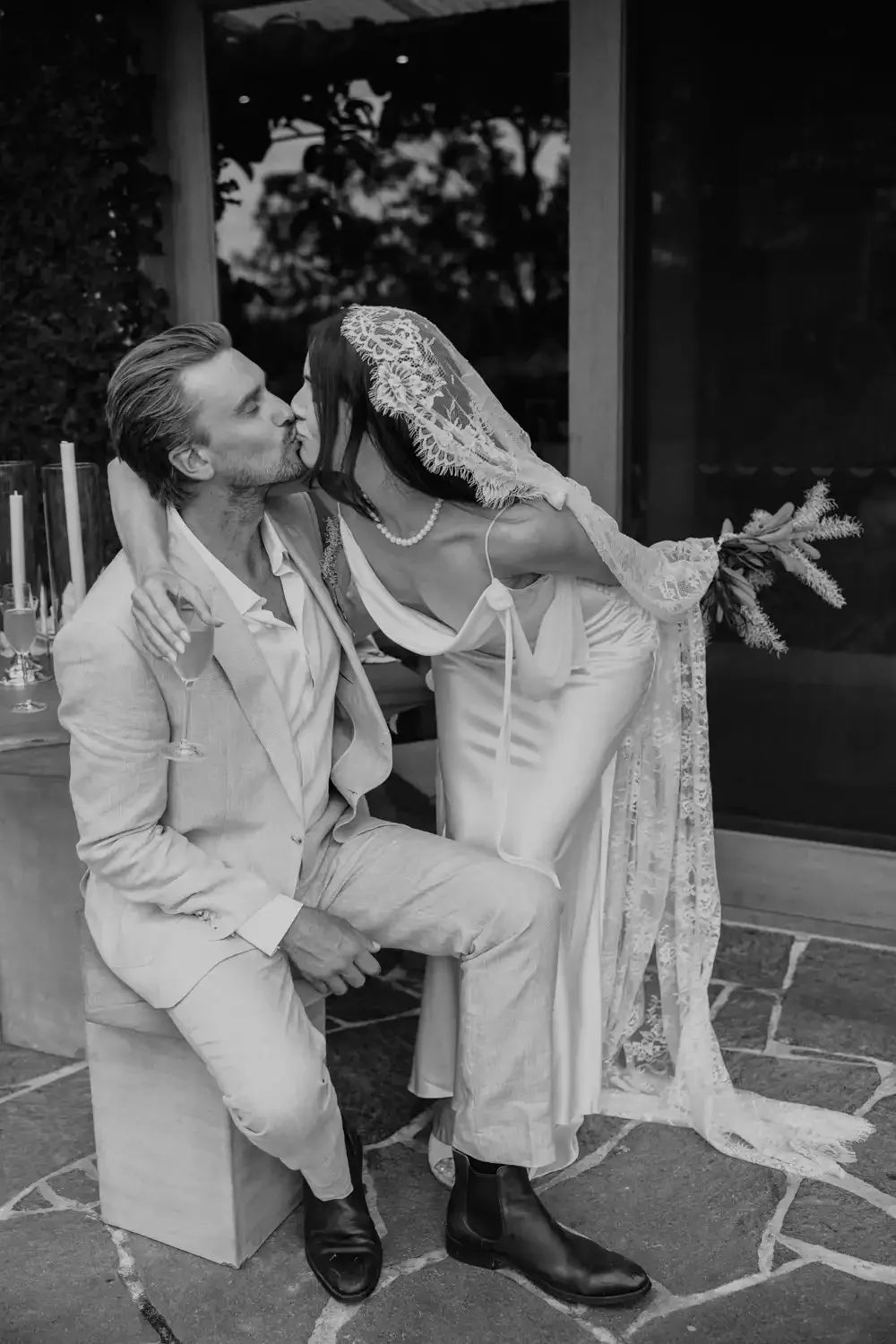 A woman in wedding dress and veil is kissing a man in a light-colored suit on a wedding day. They are outdoors near a glass door with trees in the background.