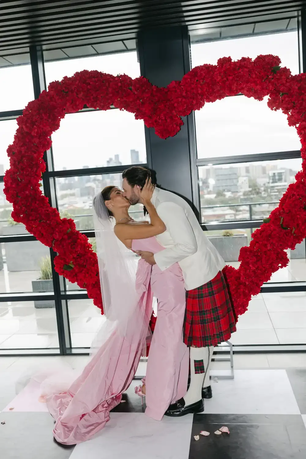A bride and groom kissing inside a heart-shaped flower arrangement at their wedding celebration.