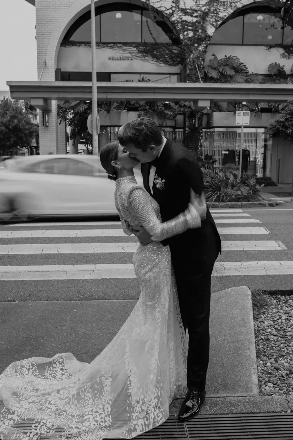 A black and white photo of a newlywed couple sharing a kiss on a city sidewalk, with a blurred car passing by in the background.