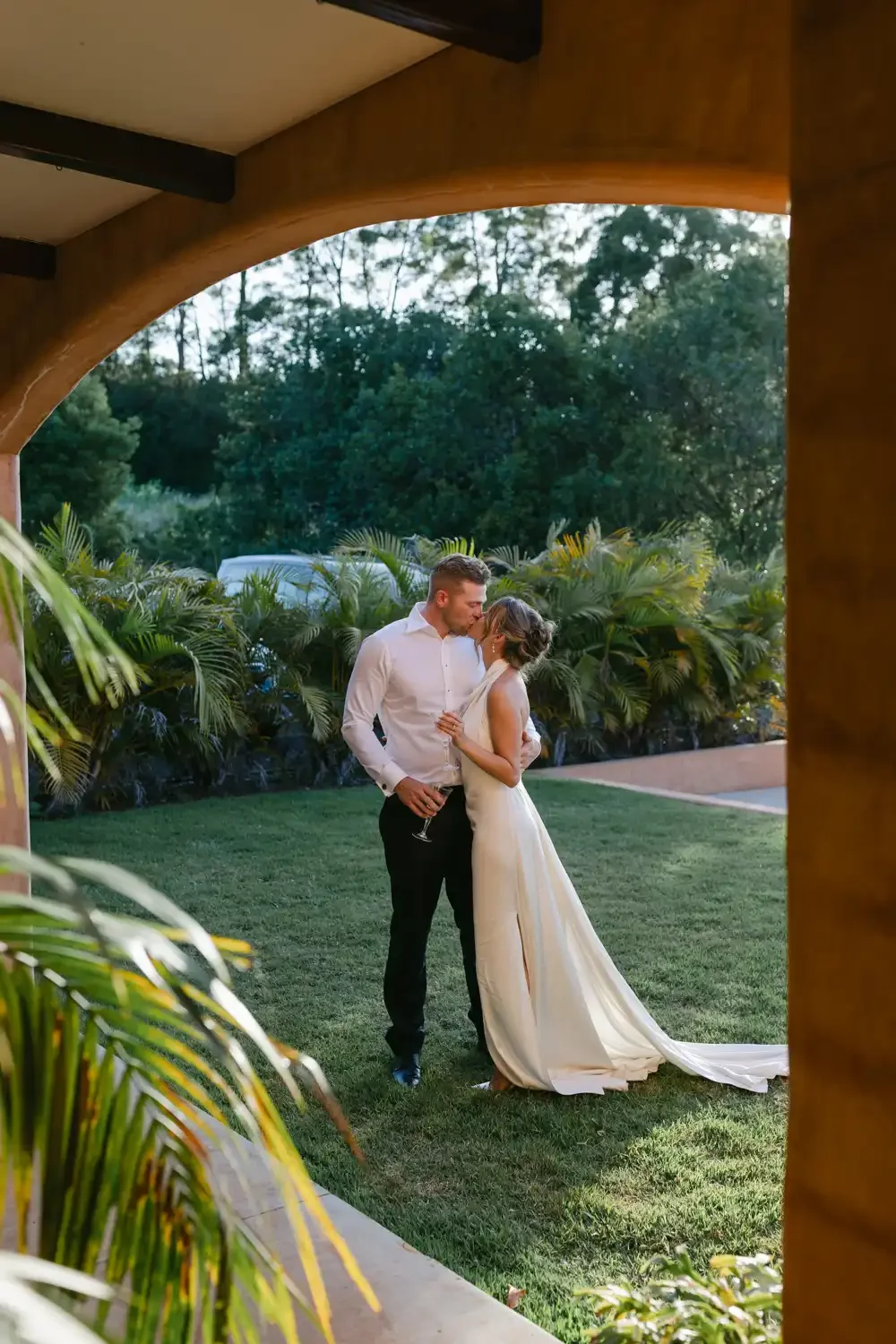 A couple in wedding attire sharing a kiss outdoors, framed by an archway made of wood with lush greenery in the background.