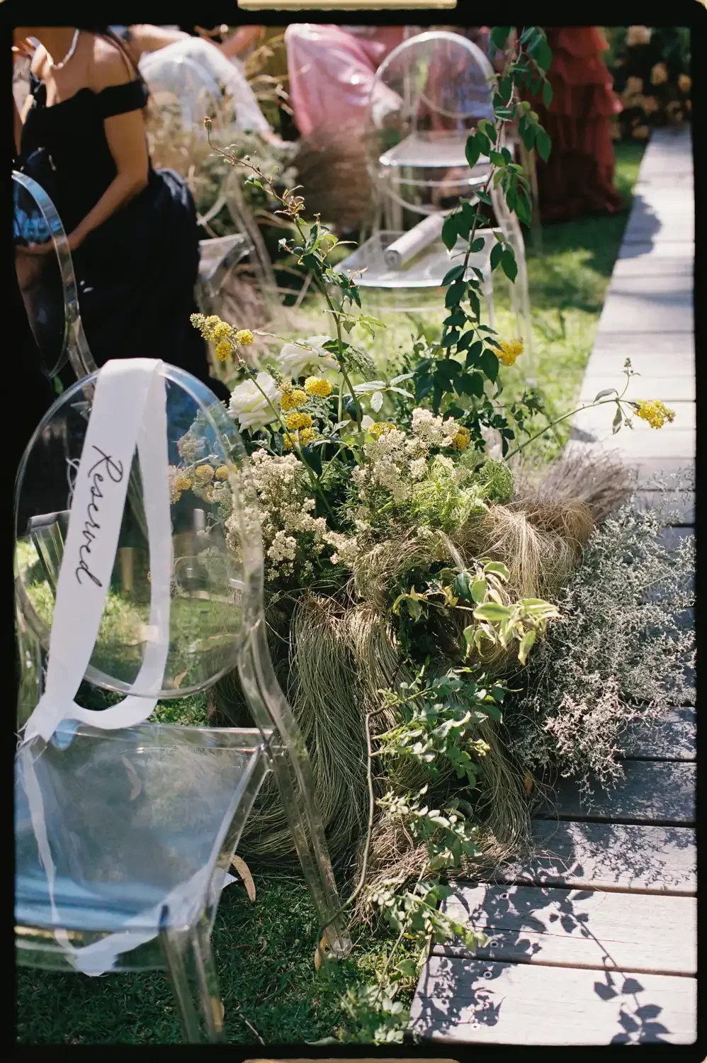 A wedding or event setup with clear acrylic chairs decorated with white ribbons, arranged on a grassy area alongside a flower and greenery arrangement on a wooden pathway.