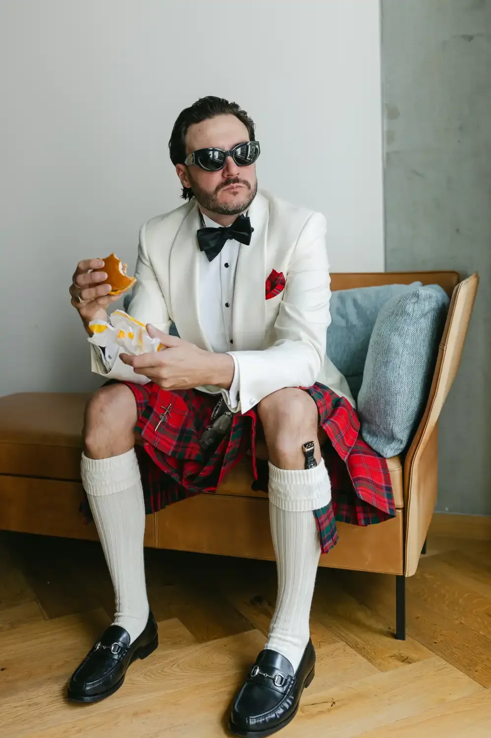 Man dressed in a tuxedo jacket, plaid kilt, knee-high socks, and dress shoes, seated on a modern couch, wearing sunglasses and eating a burger.