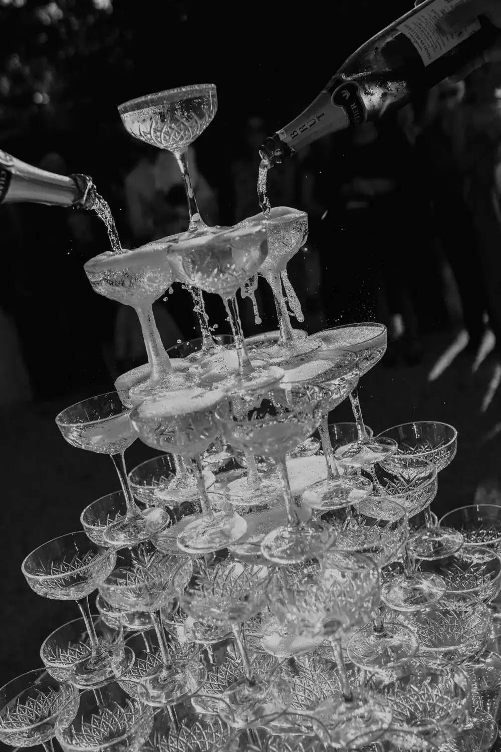 A tower of champagne glasses is being filled with champagne from bottles, and the glasses are cascading with liquid as they overflow.