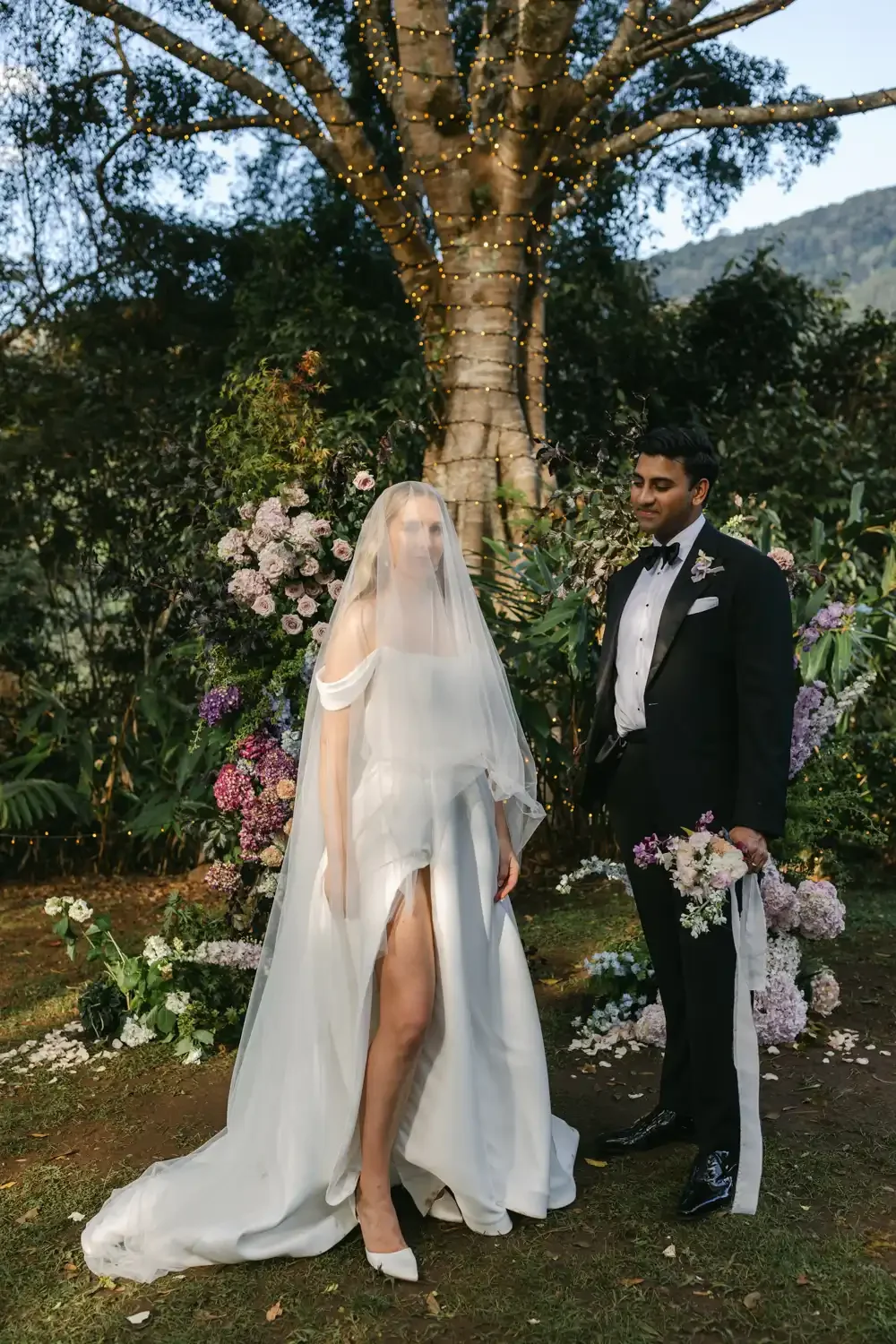 A bride and groom standing in a garden with a large tree decorated with string lights behind them. The bride is wearing a white wedding dress and veil, holding a bouquet of flowers, with her dress revealing one leg. The groom is in a black tuxedo wit
