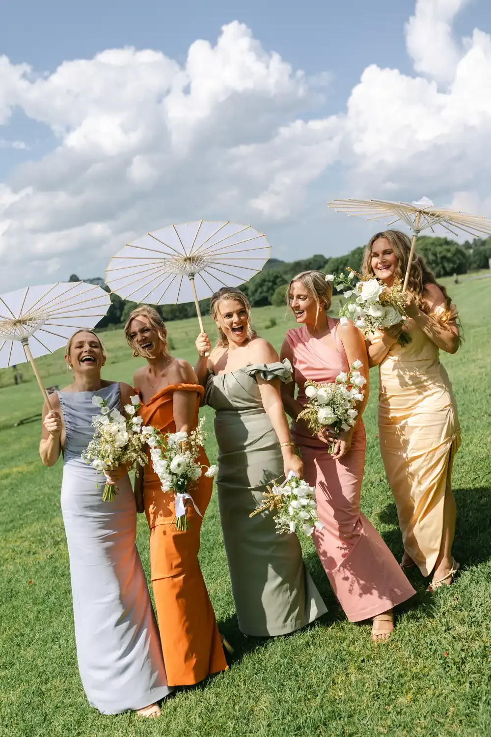 Group of six women in colorful dresses holding bouquets of flowers and parasols, smiling and walking outdoors on a sunny day with green hills and clouds in the background.