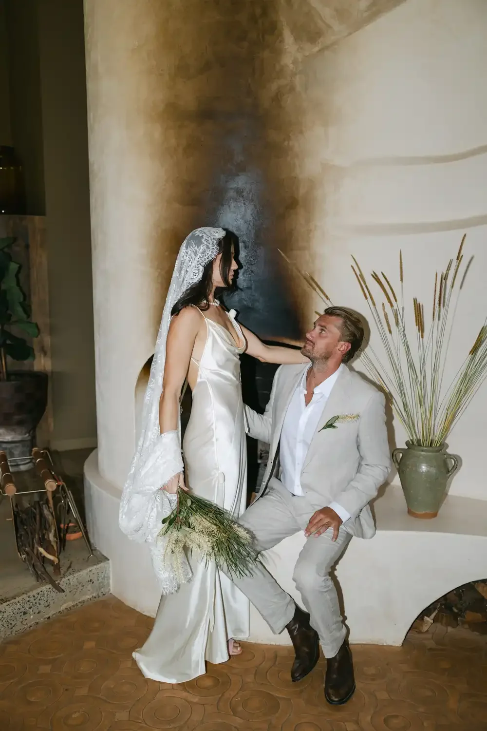 A couple dressed in wedding attire, with the woman holding a bouquet, sitting inside a warmly decorated room. The woman wears a satin wedding gown and veil, standing beside the man dressed in a light-colored suit, who is sitting on a ledge with plant