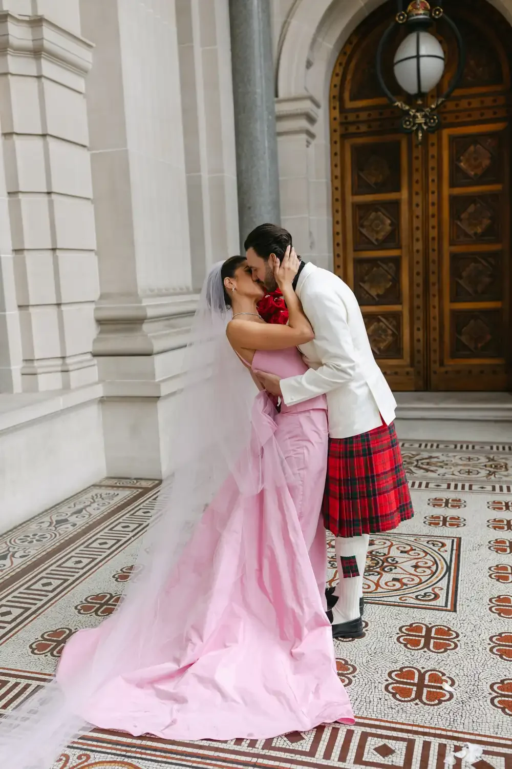 Bride and groom sharing a kiss in front of a wooden door inside a historic building, with ornate tiled floor and stone walls. The bride is wearing a pink dress with a veil, and the groom is in a white jacket with a red tartan kilt.