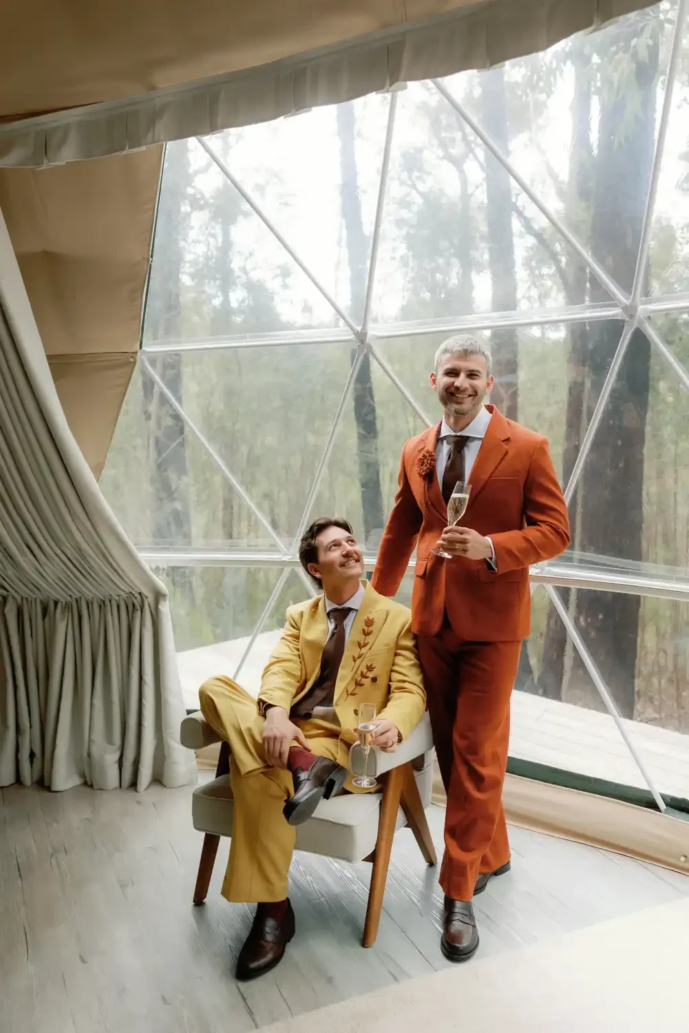 Two men in colorful suits celebrating with champagne inside a modern geodesic dome with large glass windows overlooking a forest.