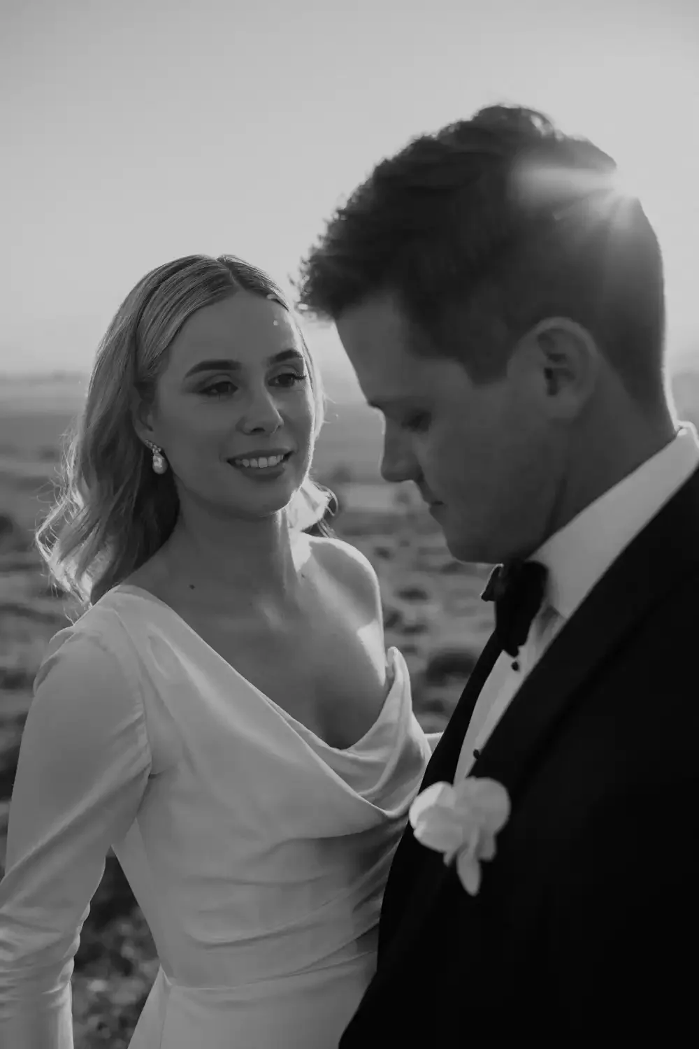 Black and white photo of a bride and groom outdoors, with the bride smiling and looking at the groom, who is looking down, against a sunset background.