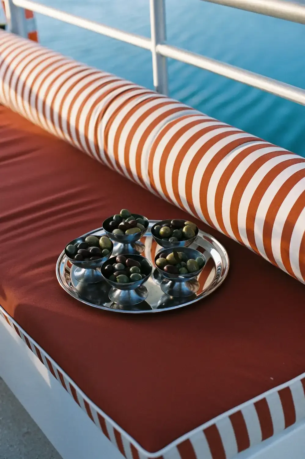 Tray with five bowls of green and dark olives on a deck with a striped orange and white railing and a blue ocean background.
