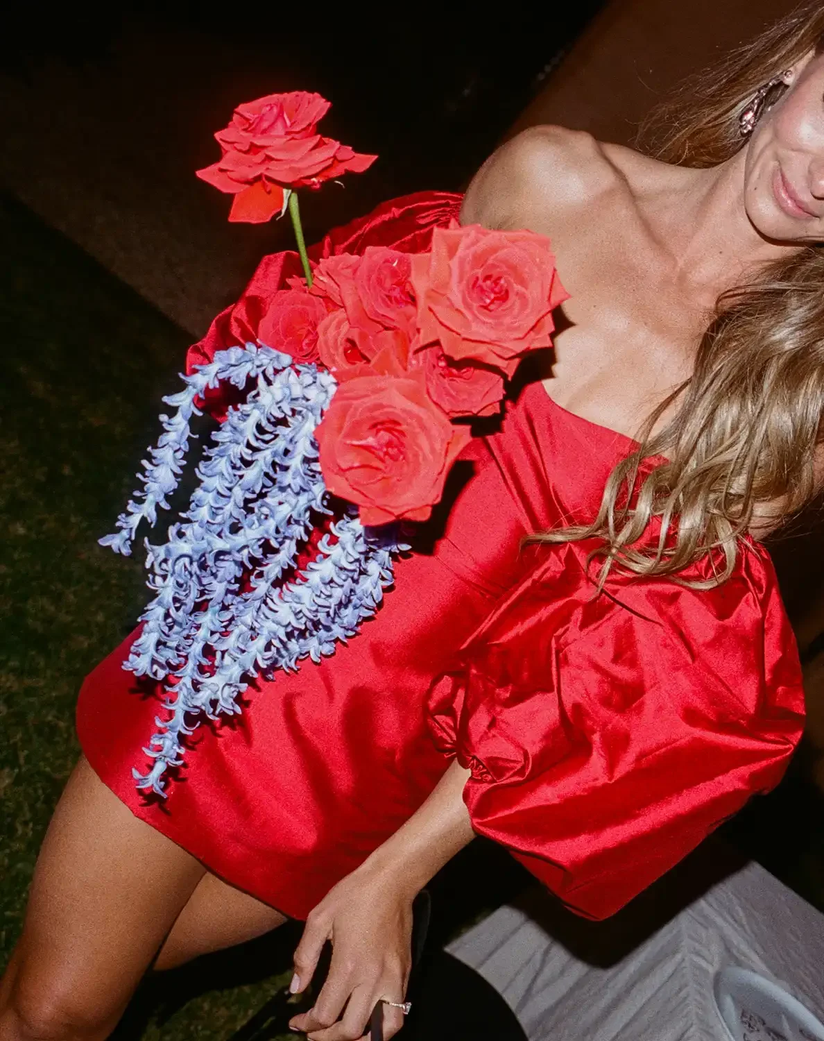 Woman wearing a red dress adorned with red roses and blue orchids, with earrings and a ring, partially visible in the photo.