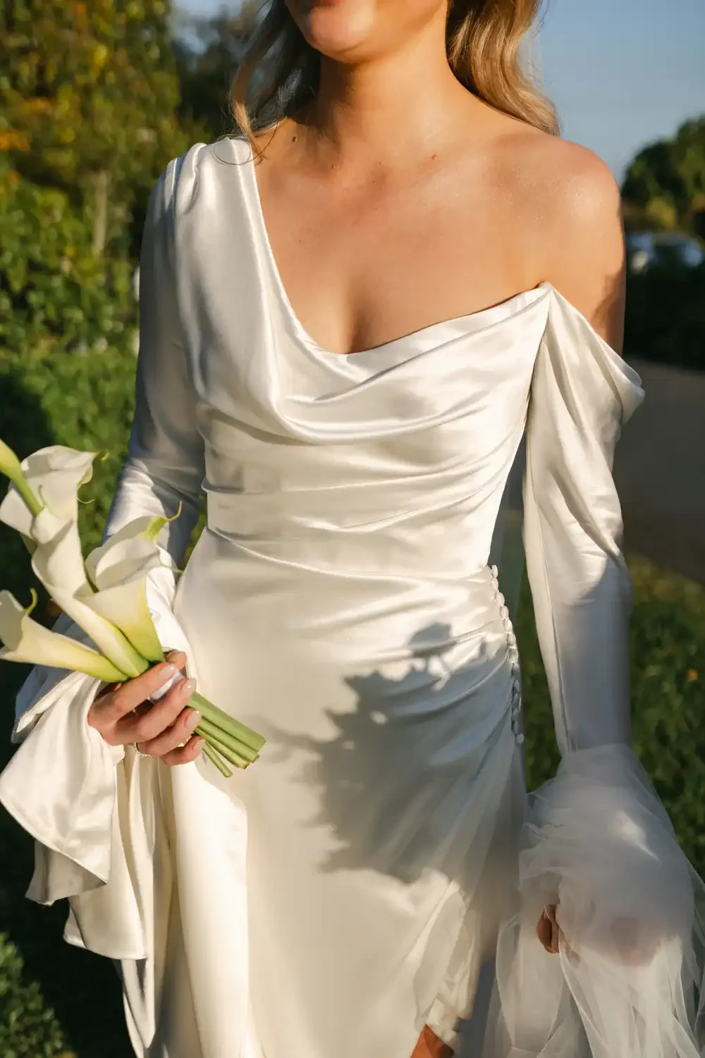 A woman in a white satin wedding dress holding a bouquet of calla lilies outdoors.