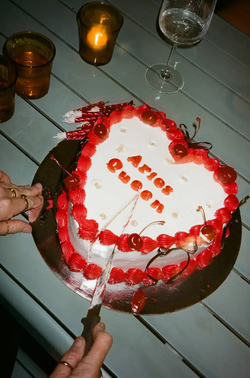 A heart-shaped birthday cake with white icing, red frosting border, cherries on top, and the message 'Arielle Duran' written with red letters. The cake is being cut by a person holding a knife. There are drinks and lit candles on the table around the