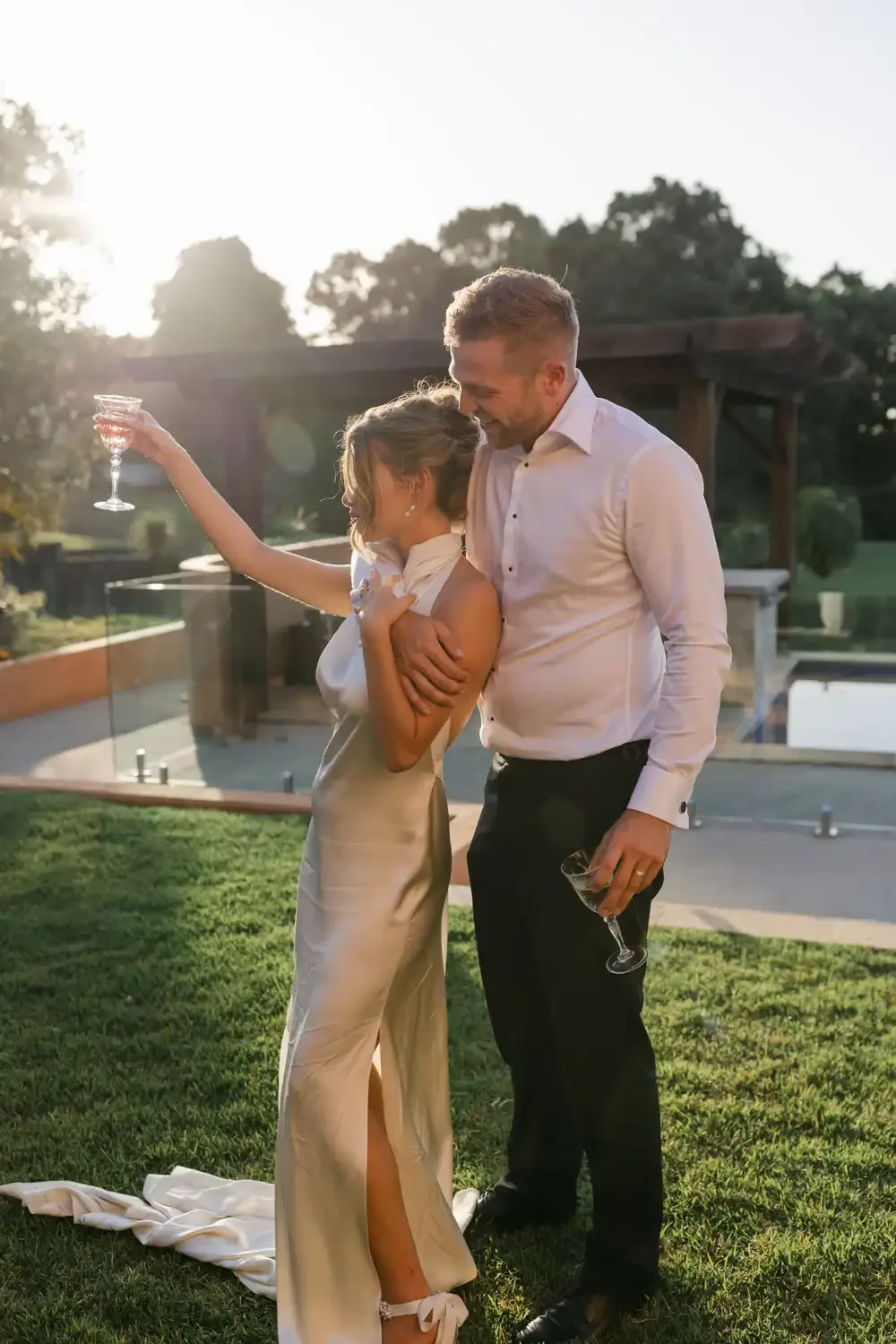 A couple celebrating outdoors in the evening, with the woman holding a glass of wine, and both smiling and looking at each other, with a backyard and trees in the background.