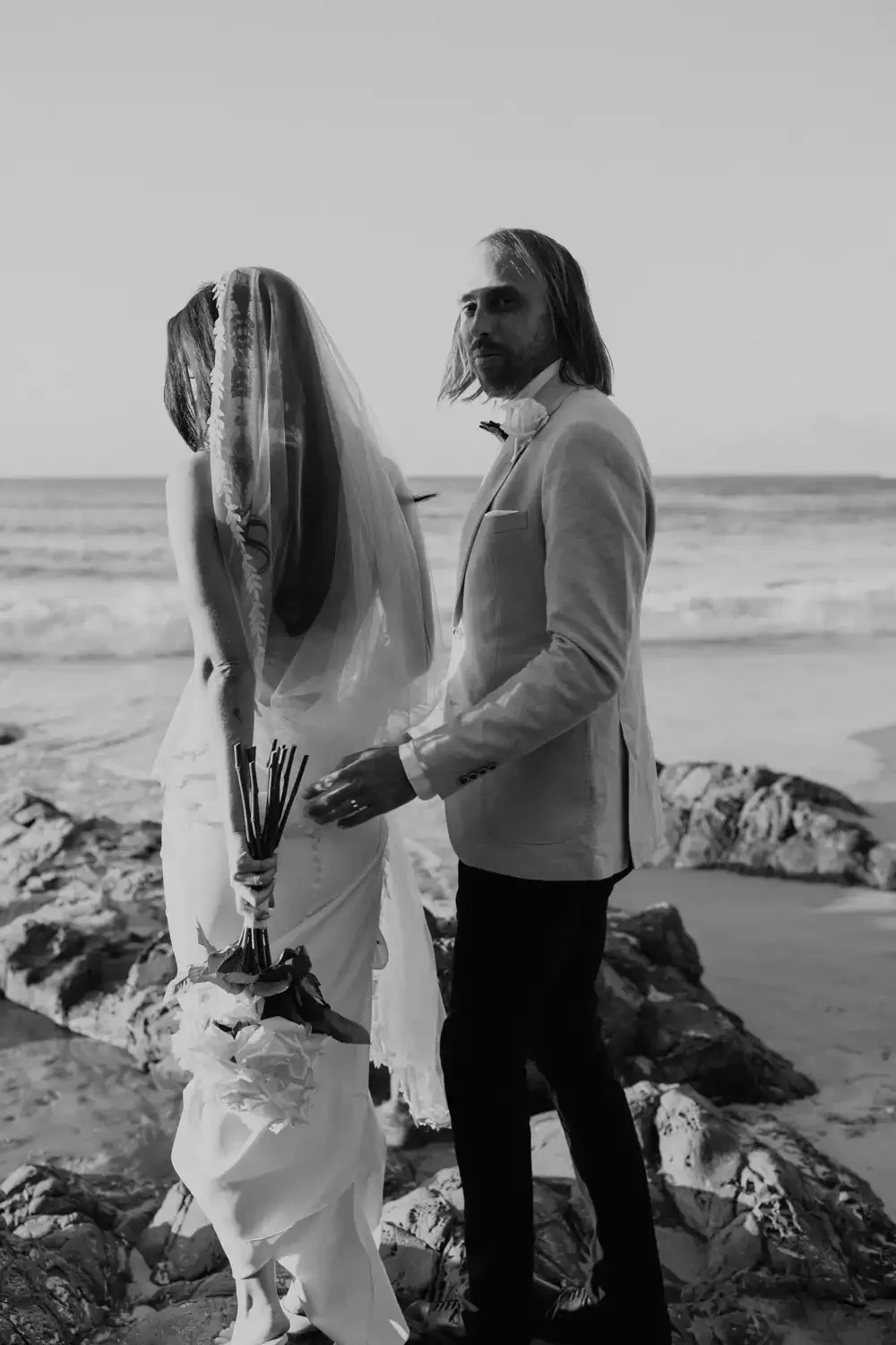 A couple dressed in wedding attire standing on a rocky beach, with the ocean in the background, during sunset, in black and white.