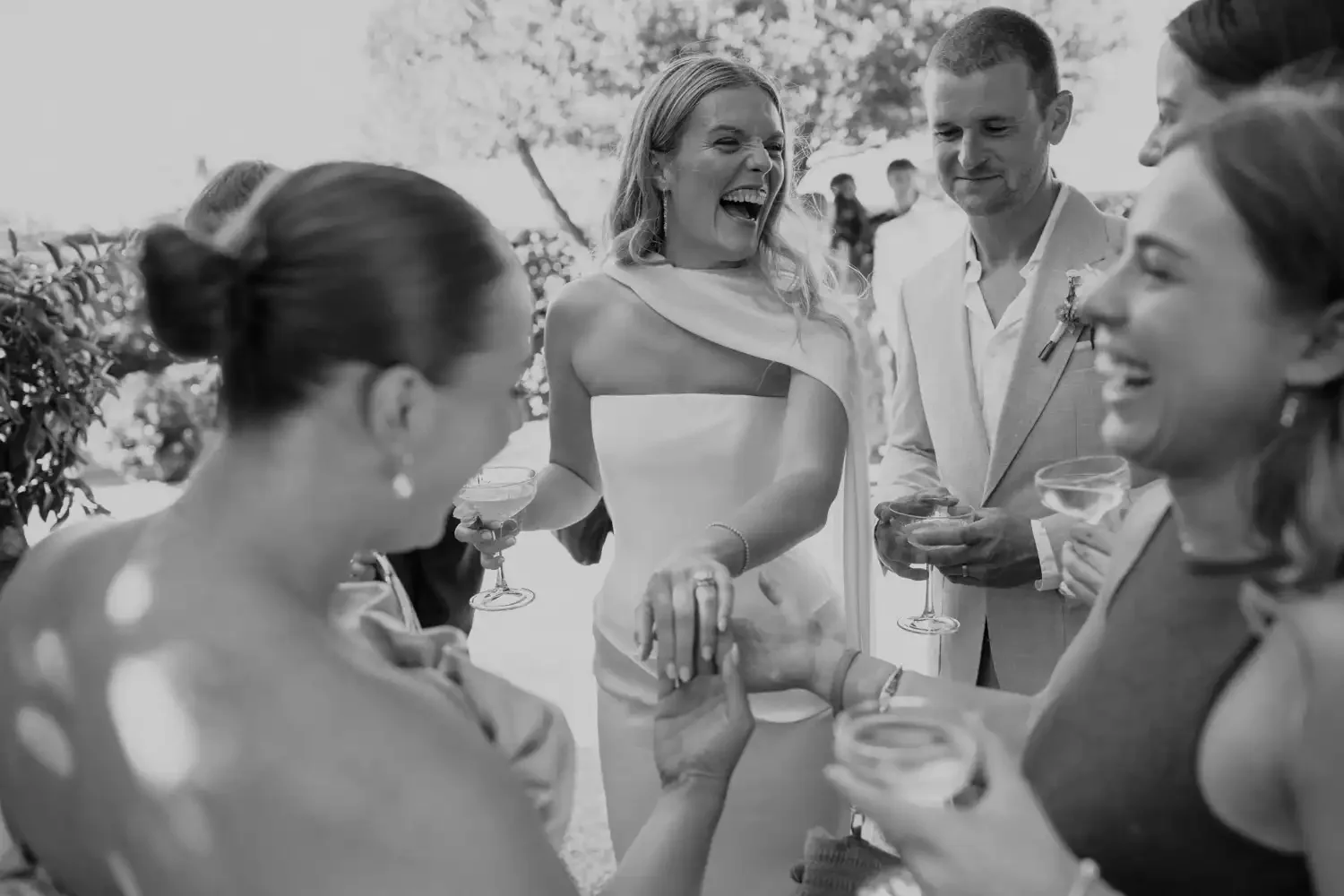 A group of five people at a wedding reception, smiling and laughing while holding drinks. The scene appears joyful and celebratory.