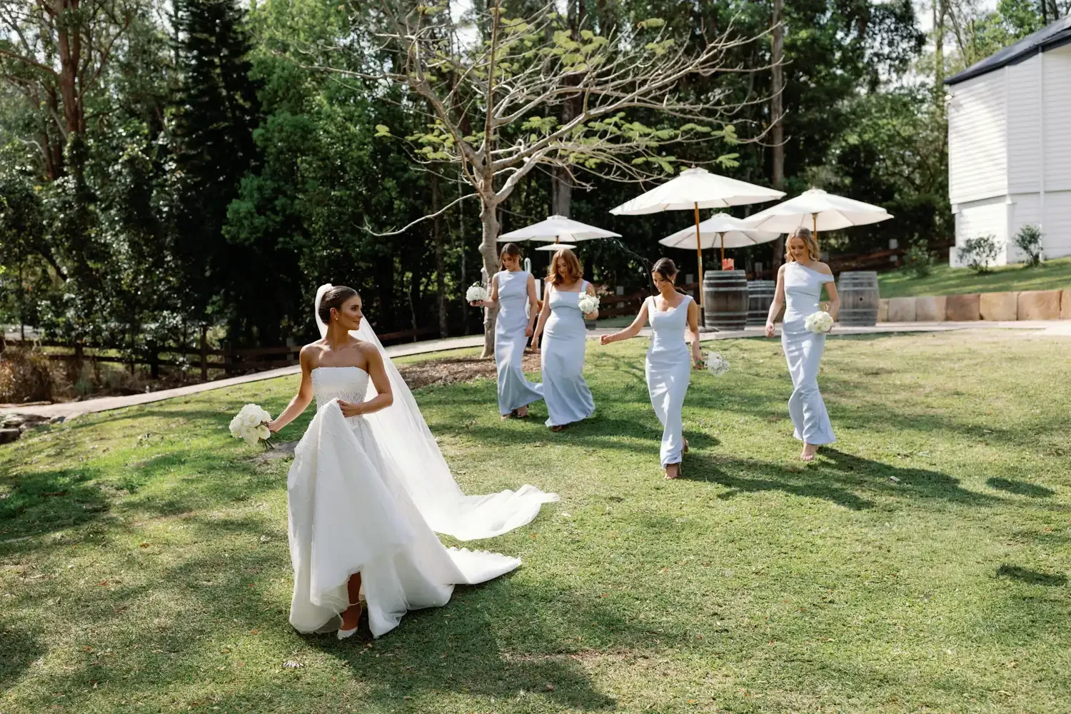 A bride in a white wedding gown walking on a grassy area holding a bouquet, while five women in light blue dresses follow behind her in an outdoor setting with trees, umbrellas, and barrels.