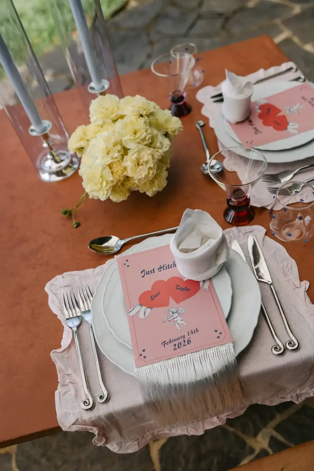 A decorated table with a pink and white theme, featuring a floral centerpiece, vintage-style silverware, pink-tinted glasses, and a Valentine's Day-themed menu card with the date February 14th, 2026, indicating a romantic occasion.