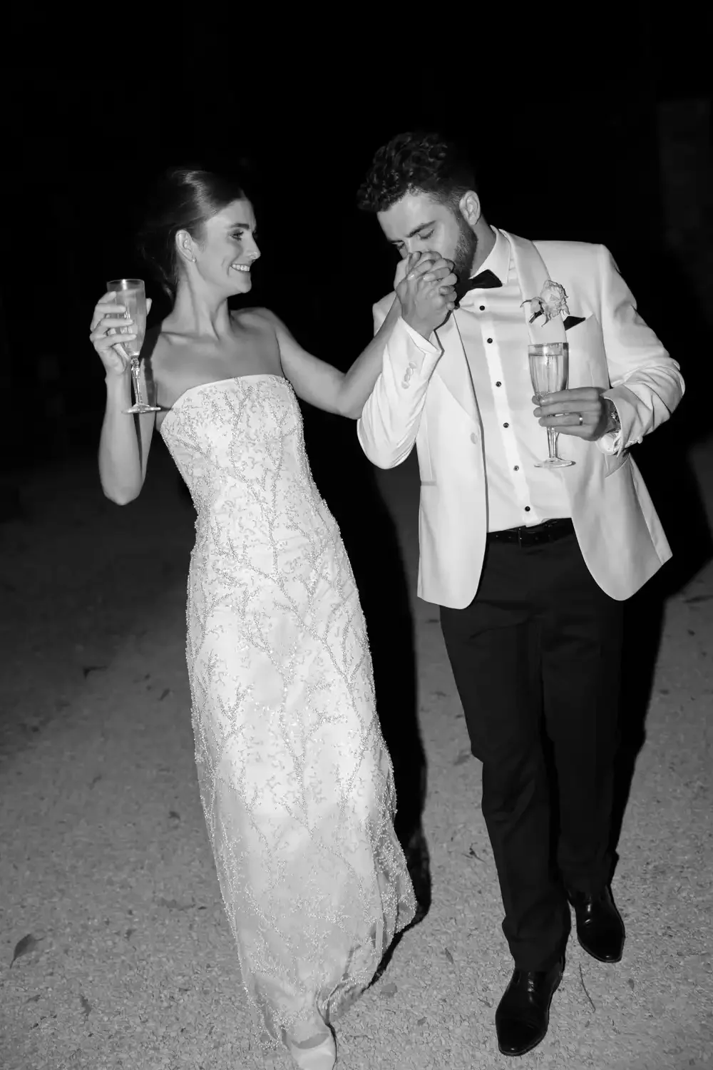 A bride and groom at their wedding reception, with the groom kissing the bride's hand as she holds a glass of champagne. The bride is in a strapless wedding gown, and the groom is dressed in a tuxedo with a boutonniere.