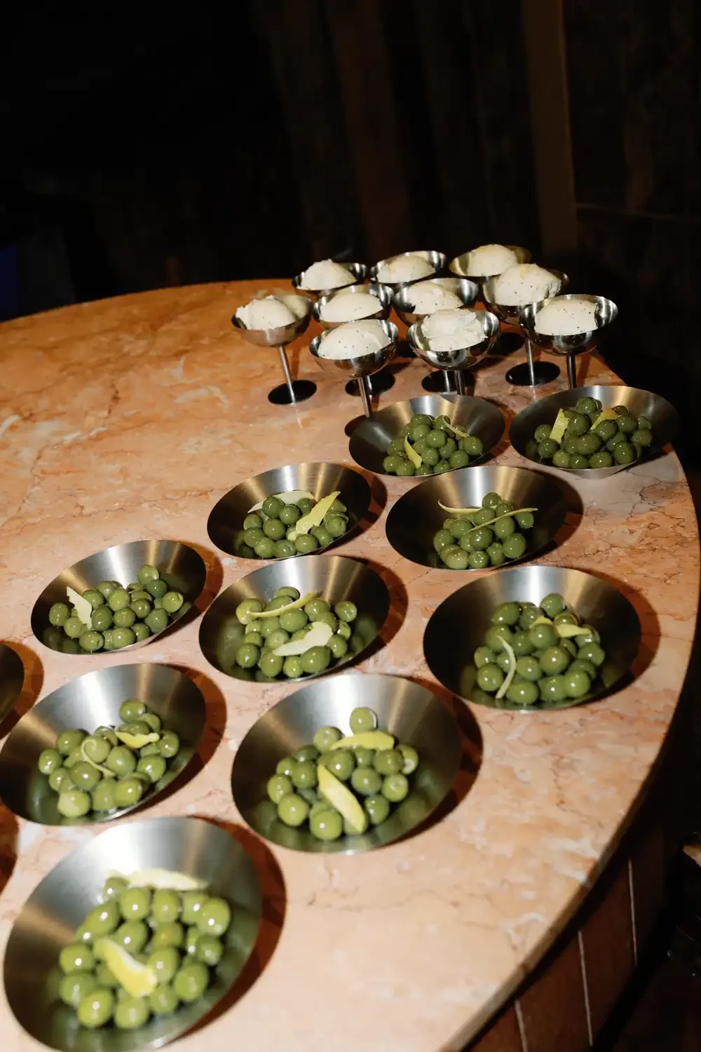 Assorted appetizers on a marble table, including small metal bowls of green olives with lemon slices and skewered cheese or meat bites.