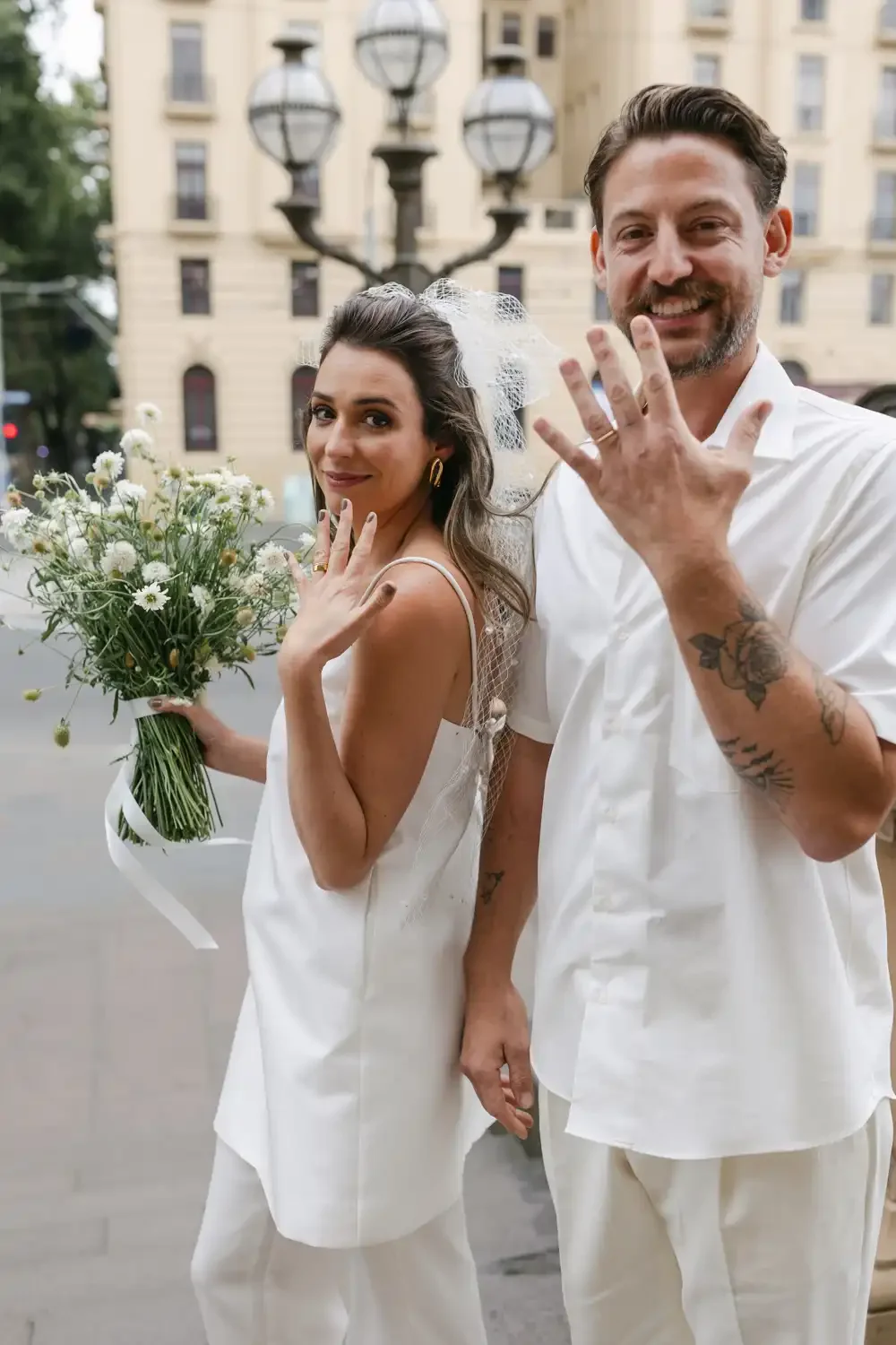 A bride and groom showing off their wedding rings outside in an urban setting, with the bride holding a bouquet of white daisies and the groom displaying tattoos on his arm, both smiling.