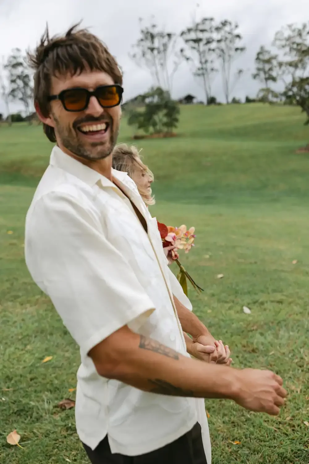 A smiling man with sunglasses and a beard holding hands with a woman with floral bouquet on a green, grassy field, trees in background.