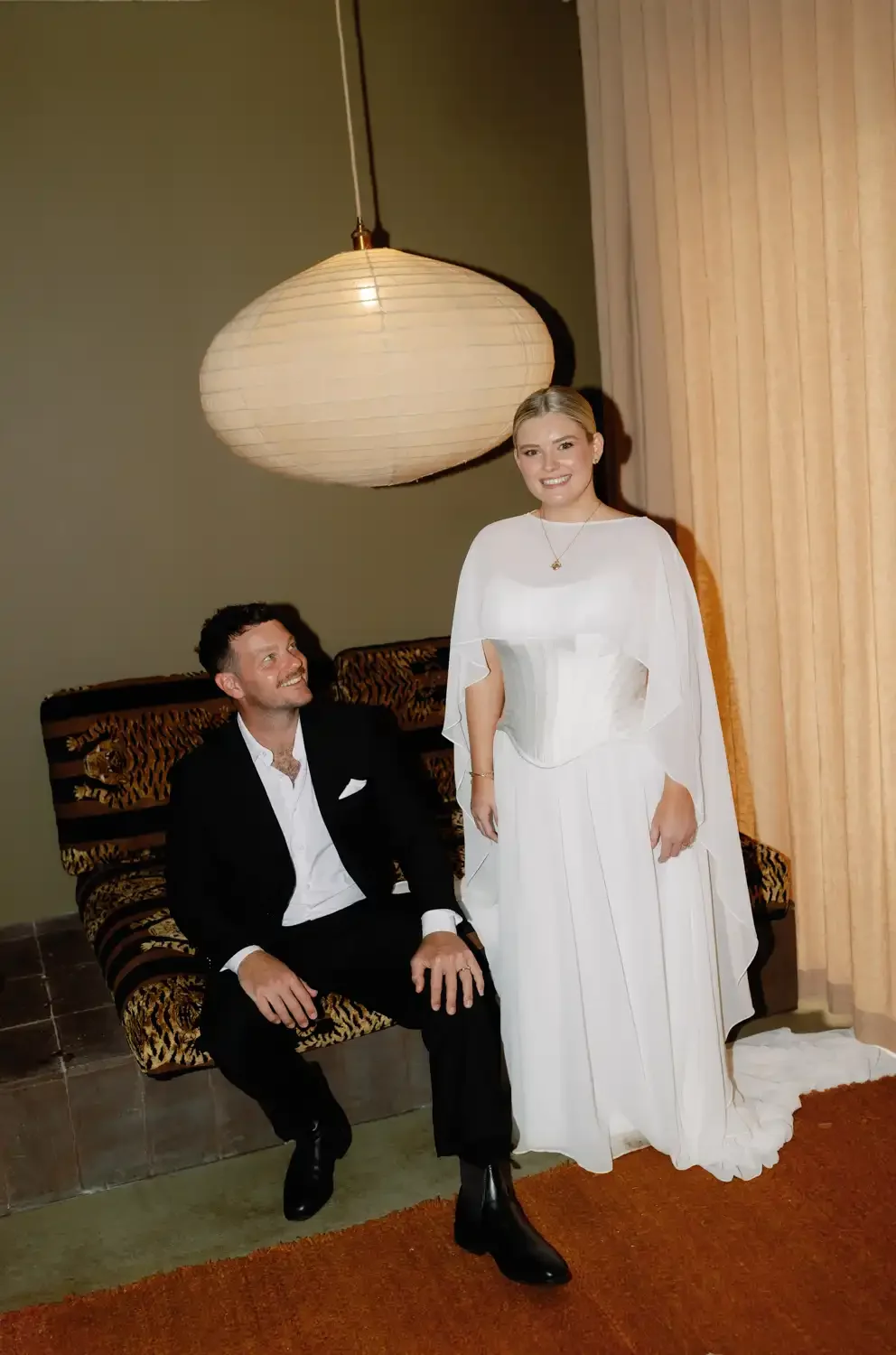 A man in a black tuxedo sitting on a patterned chair, looking up and smiling at a woman in a white dress with a corset, standing beside him and smiling.
