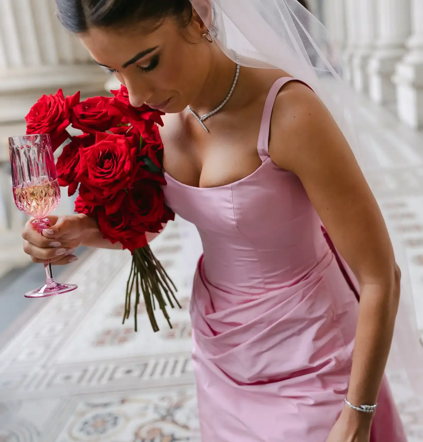 A woman in a pink dress holding a bouquet of red roses and a glass of champagne in an elegant setting.