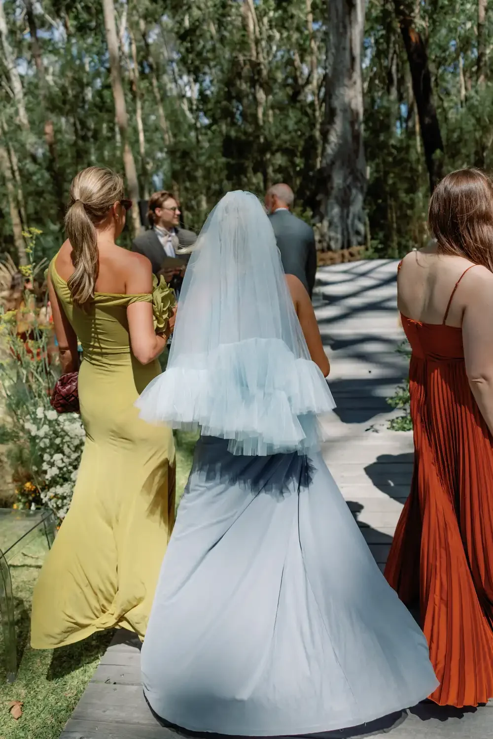 A bride wearing a white wedding dress and veil walking down the aisle at an outdoor wedding ceremony in a forest, accompanied by women in colorful dresses.
