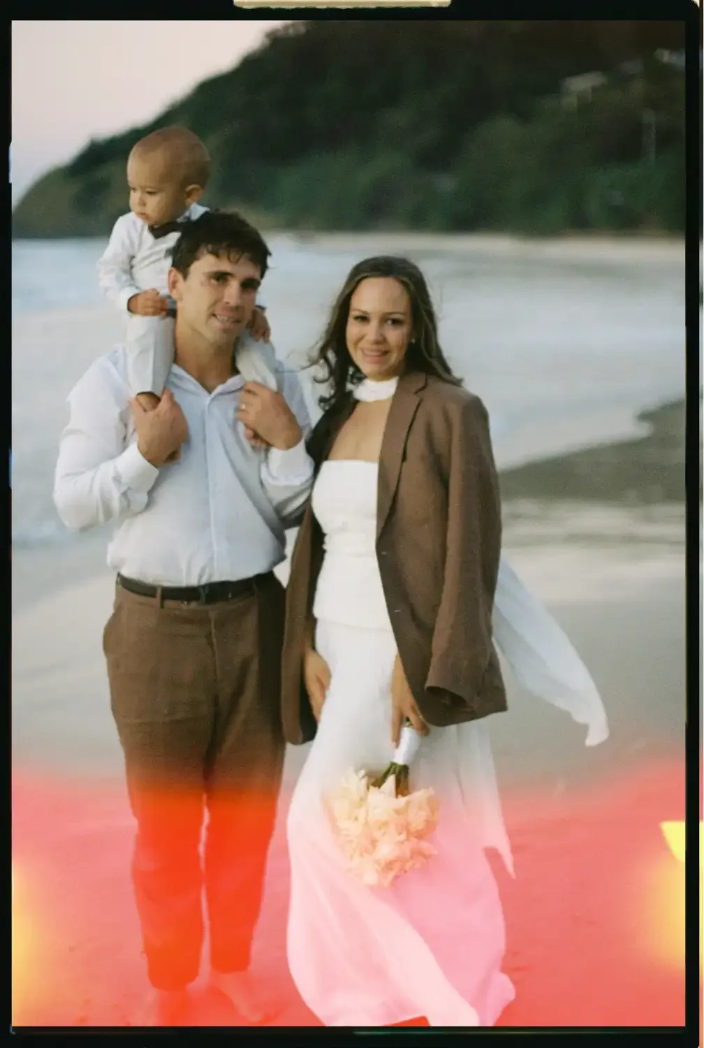 A family of three on a beach with water and green hills in the background. The man is carrying a young child on his shoulders, and the woman is standing next to them holding a bouquet of flowers.