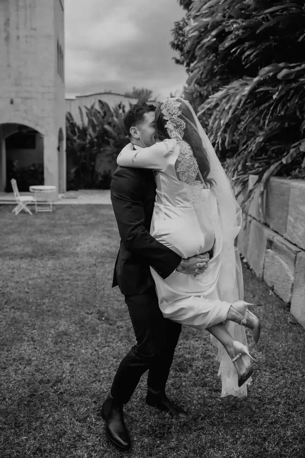 A joyful bride being lifted by a groom outdoors, both embracing closely, with trees and a building in the background.