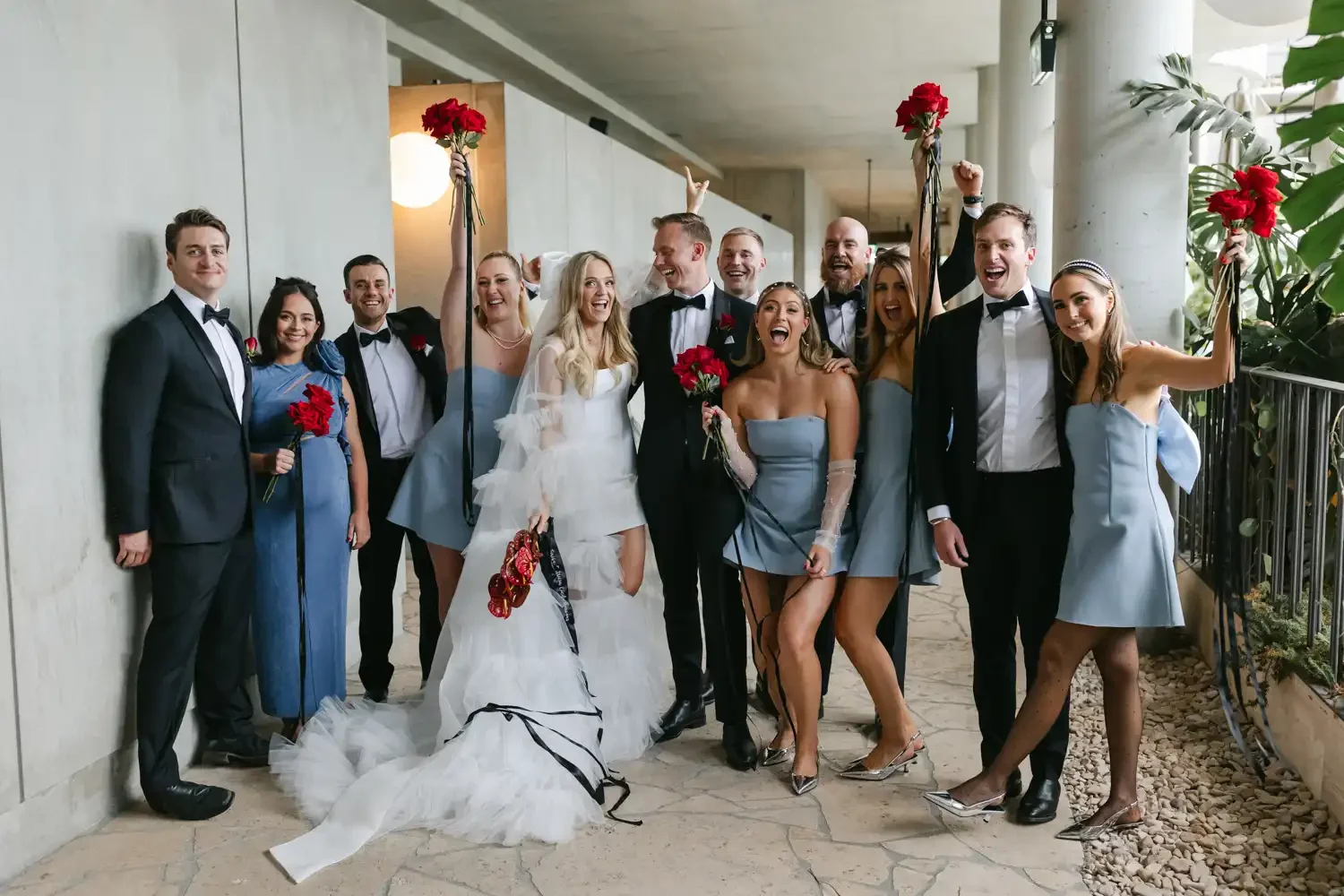 Group of people at a wedding celebration, including a bride and groom, dressed in formal attire, holding red flowers, smiling and celebrating indoors.
