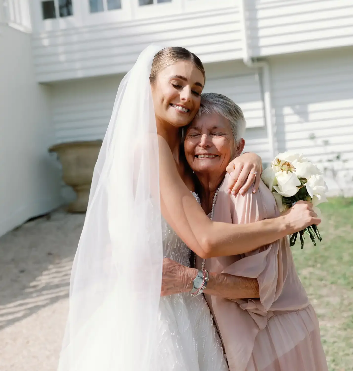 A bride in a wedding dress hugging an elderly woman who is holding a bouquet of white flowers, outside near a white building.