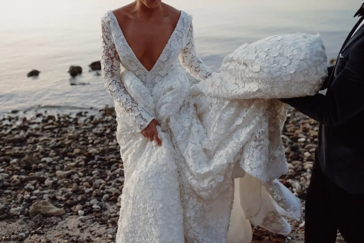 A bride in a white lace wedding dress with a deep V neckline is standing on a rocky shoreline holding her dress while an individual in dark clothing helps support it.