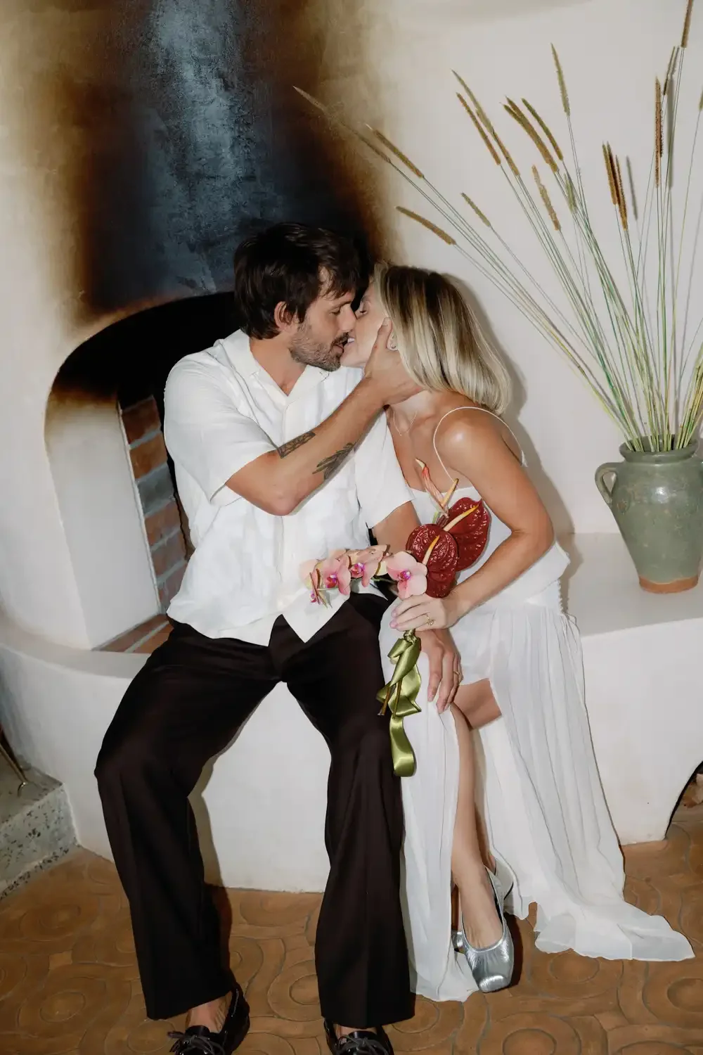 A couple shares a kiss indoors, with the woman holding a bouquet of pink orchids and red anthuriums, and the man touching her face gently. The woman is wearing a white dress with thin straps and silver high heels, while the man is dressed in a white 