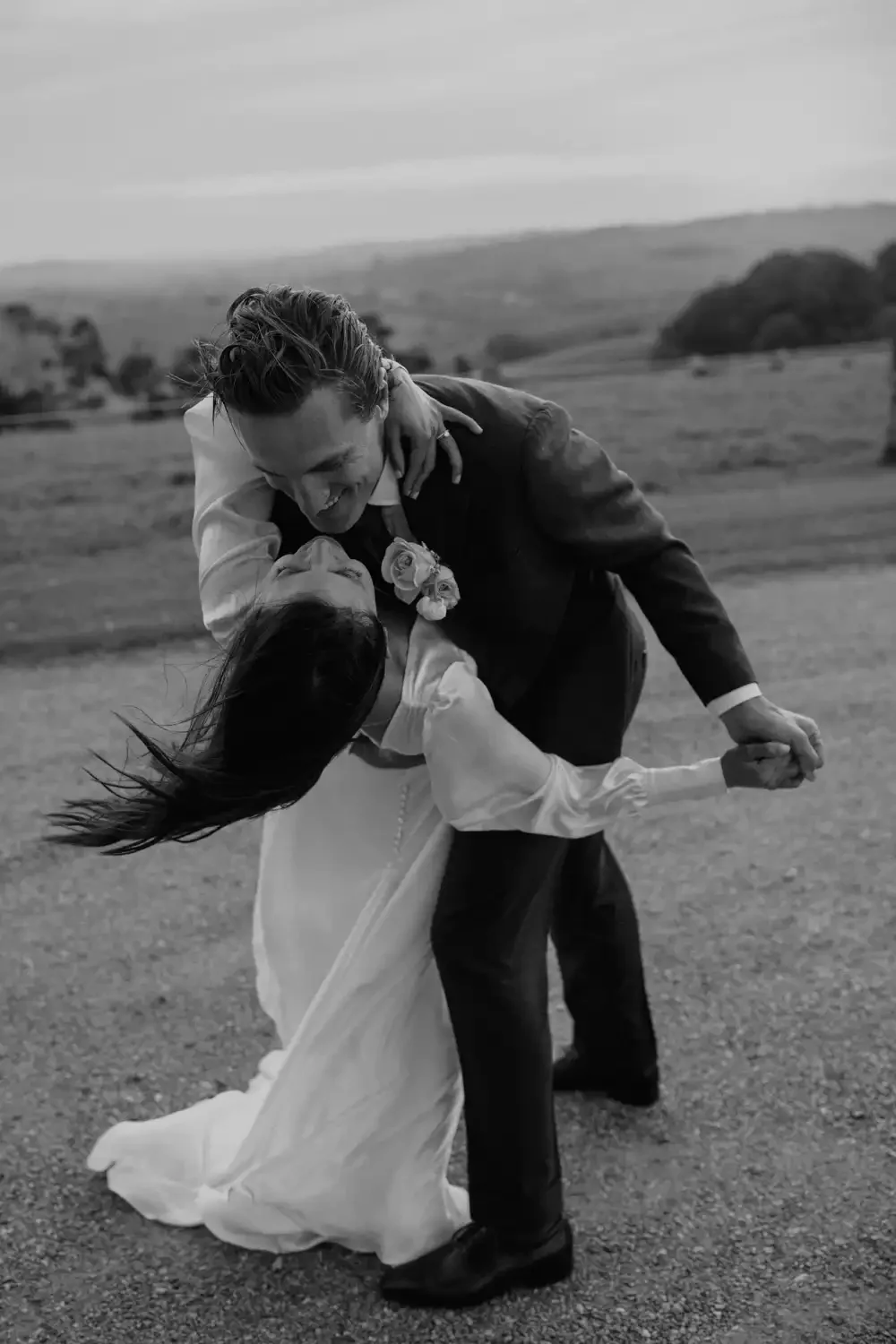 A black-and-white photo of a newlywed couple dancing outdoors in a scenic, open field, with the groom dipping the bride and both smiling.
