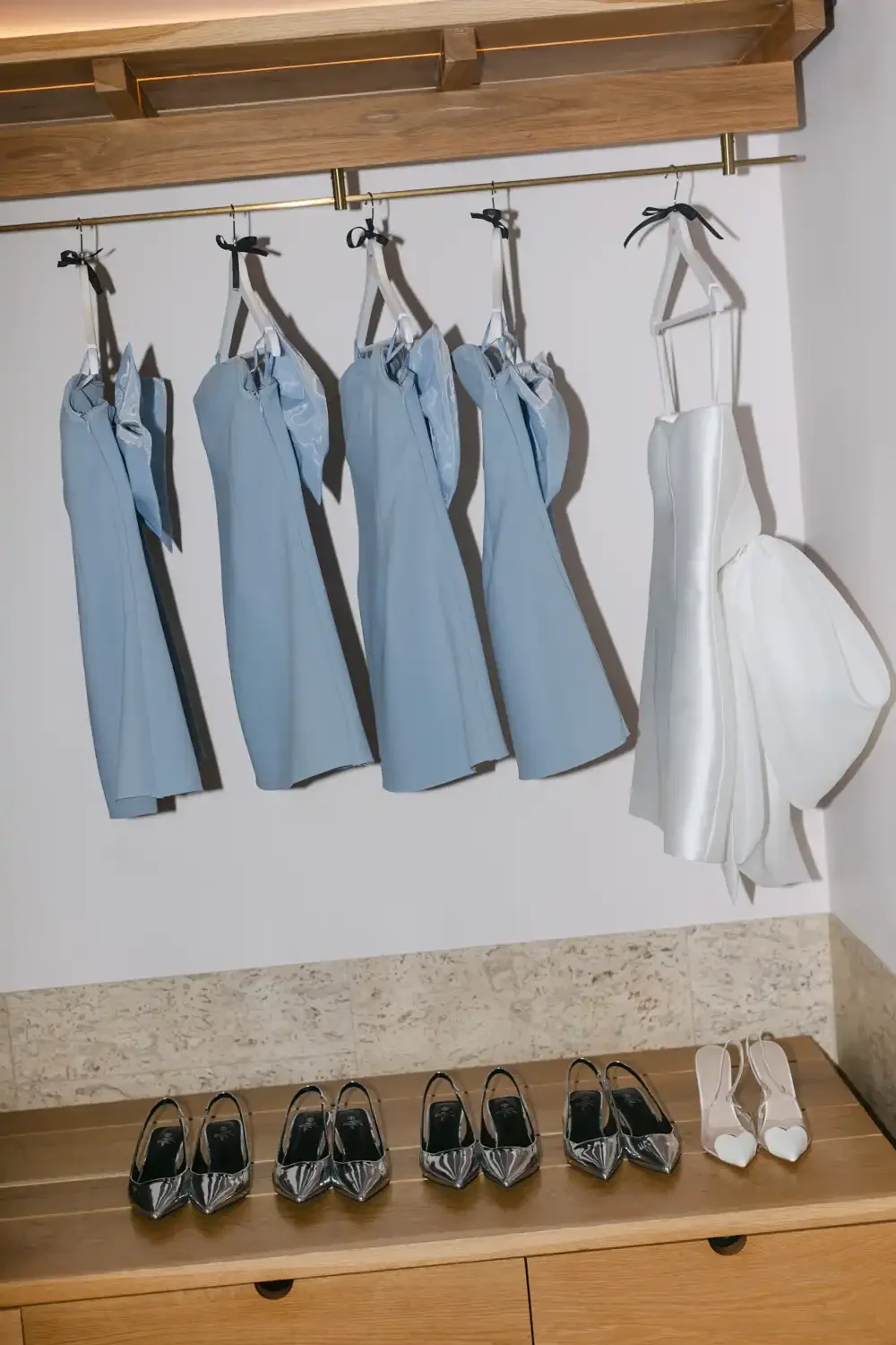 A display of five light blue dresses hanging on a gold rod, with five pairs of black and silver high heels on a wooden bench below, and a white dress with a crinoline hanging on the right side.