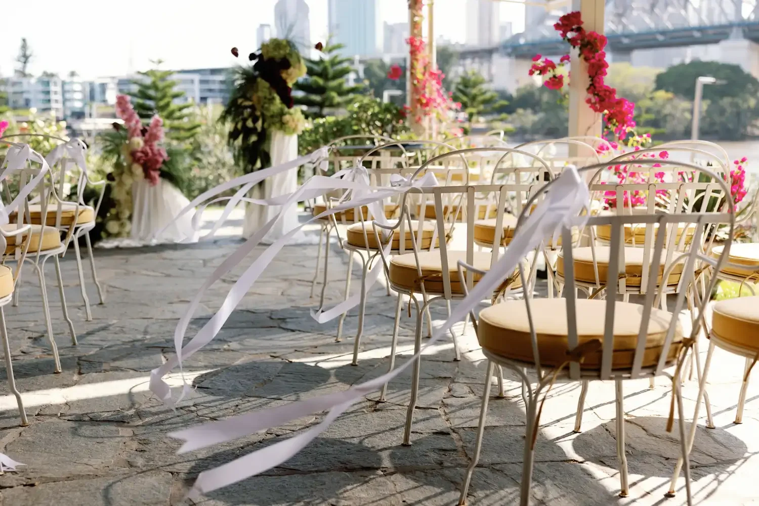 Empty outdoor wedding seating area with white chairs decorated with white ribbons, set on a stone patio with floral arrangements and city skyline in the background.
