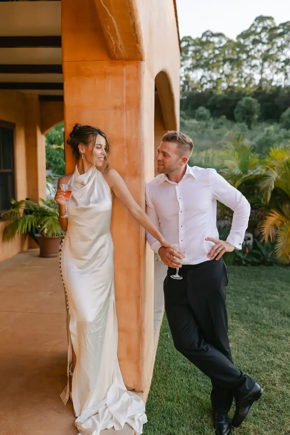 A man and woman at a social gathering, leaning against a wooden structure, holding wine glasses, smiling at each other, with tropical plants and trees in the background.
