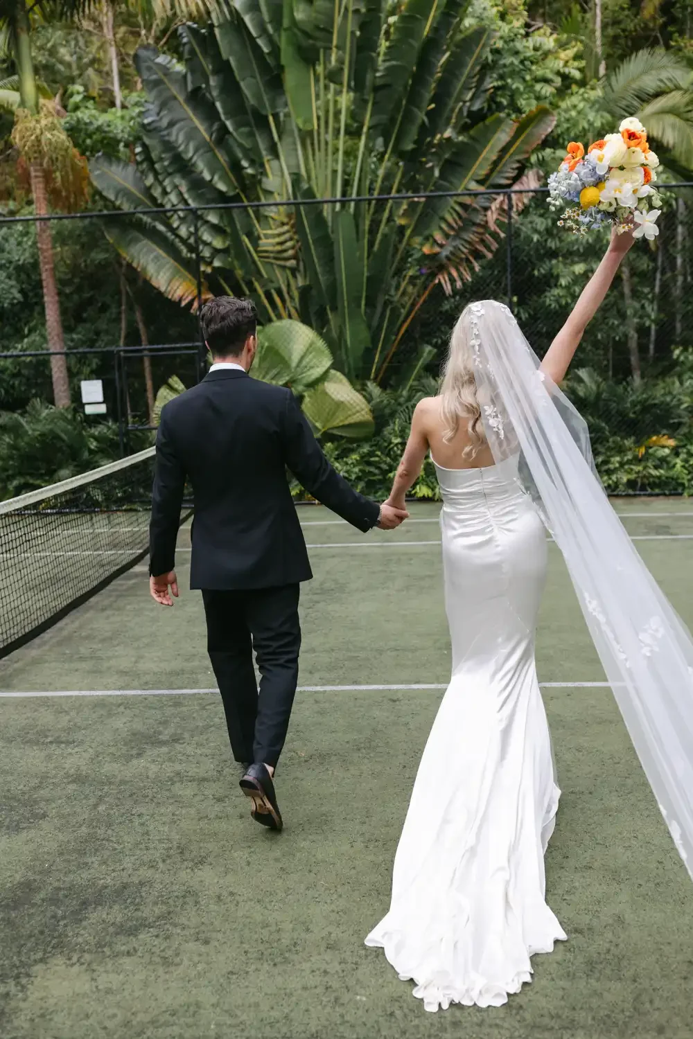 A bride and groom holding hands on a tennis court surrounded by lush greenery, with the bride holding a bouquet in one hand and wearing a white wedding dress and veil.