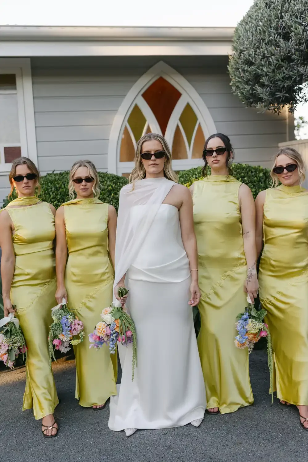 A bride and five bridesmaids standing outside in front of a house with a pointed window. The bride is in a white dress holding a bouquet, and the bridesmaids are in yellow dresses also holding bouquets. All are wearing sunglasses.