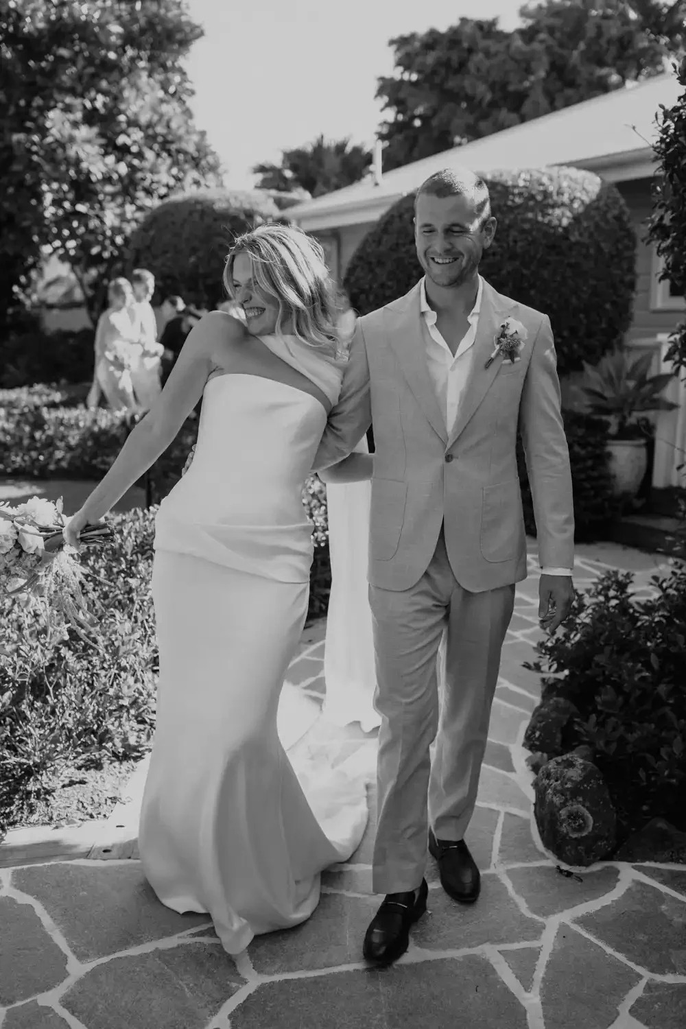 Black and white photo of a smiling bride and groom walking arm in arm outdoors during a wedding celebration. The bride is in a strapless wedding gown holding a bouquet, and the groom is in a light suit with a boutonniere.