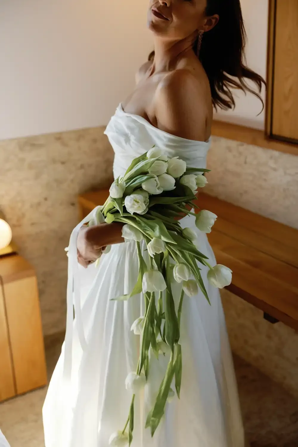 Woman in a white dress holding a bouquet of white tulips in a room with wooden furniture and a brick wall background.