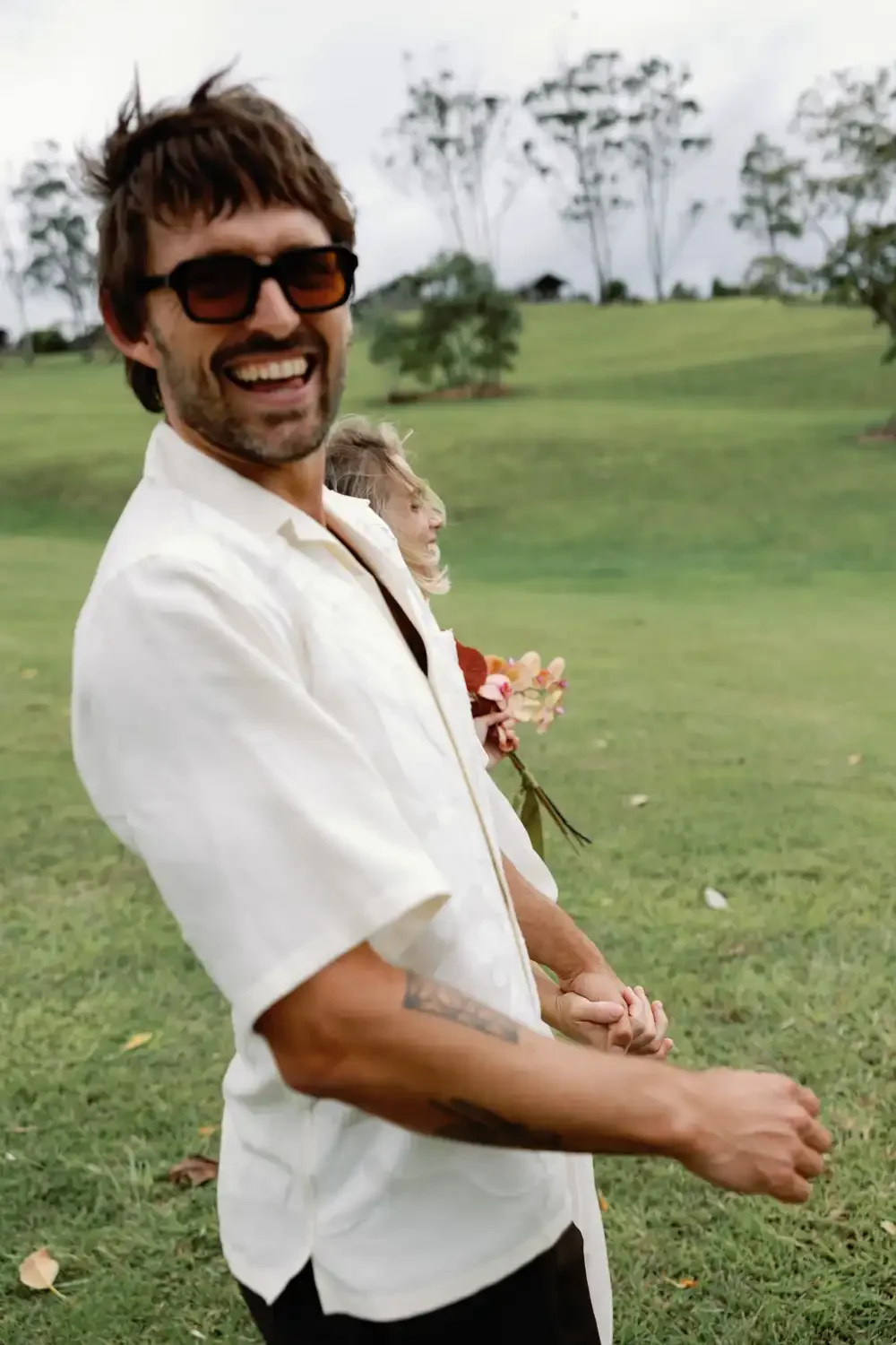 Smiling man with sunglasses holding hands with a woman and a child with a bouquet of flowers, outdoors on a grassy field.