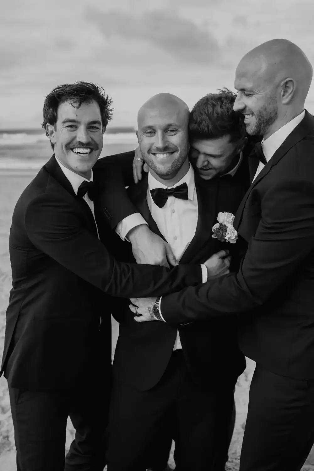 Four men in tuxedos smiling and embracing on a beach in black and white.