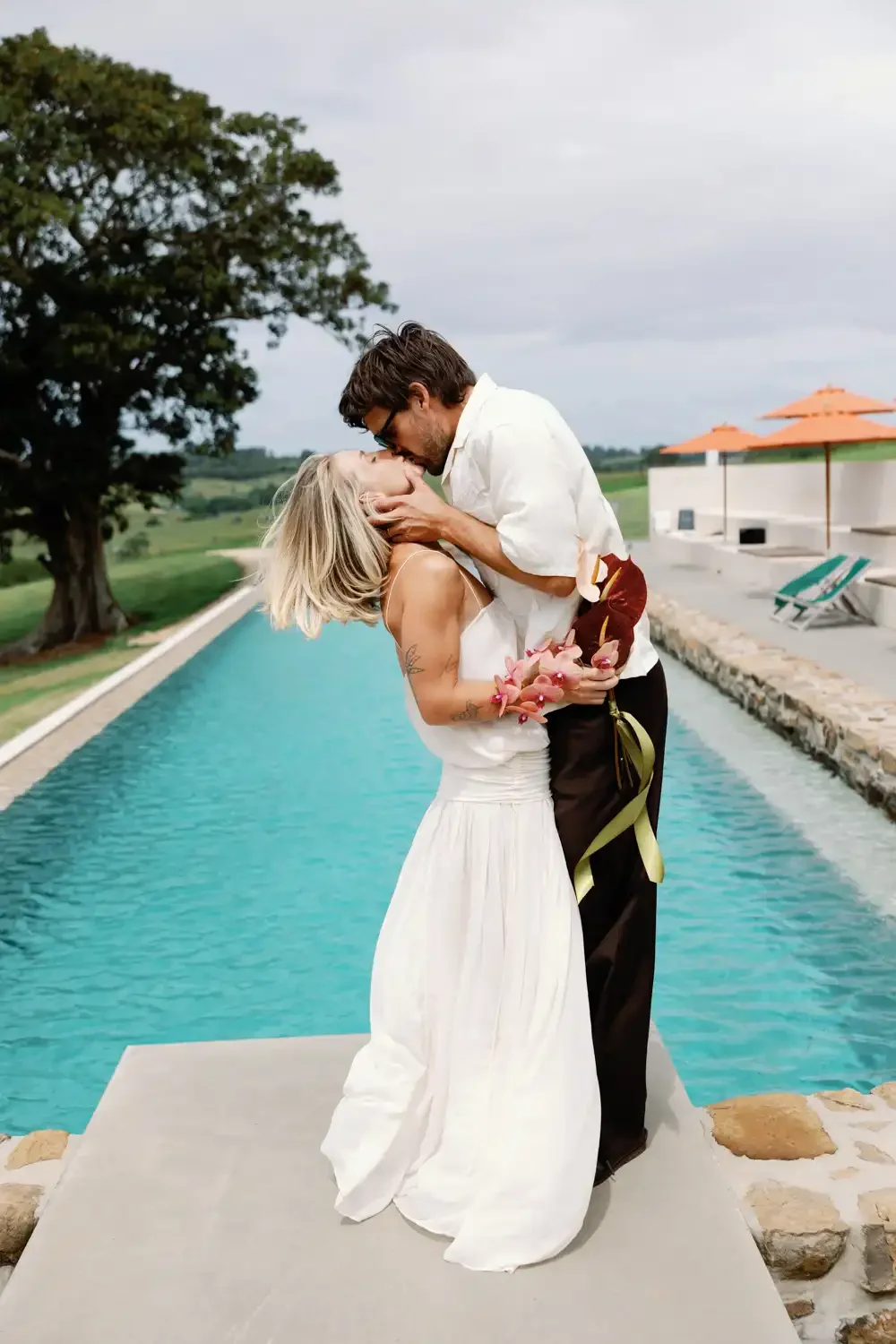 A couple sharing a romantic moment by a swimming pool, with the man lifting the woman who is holding a bouquet of pink flowers, surrounded by an outdoor setting with umbrellas and lounge chairs.