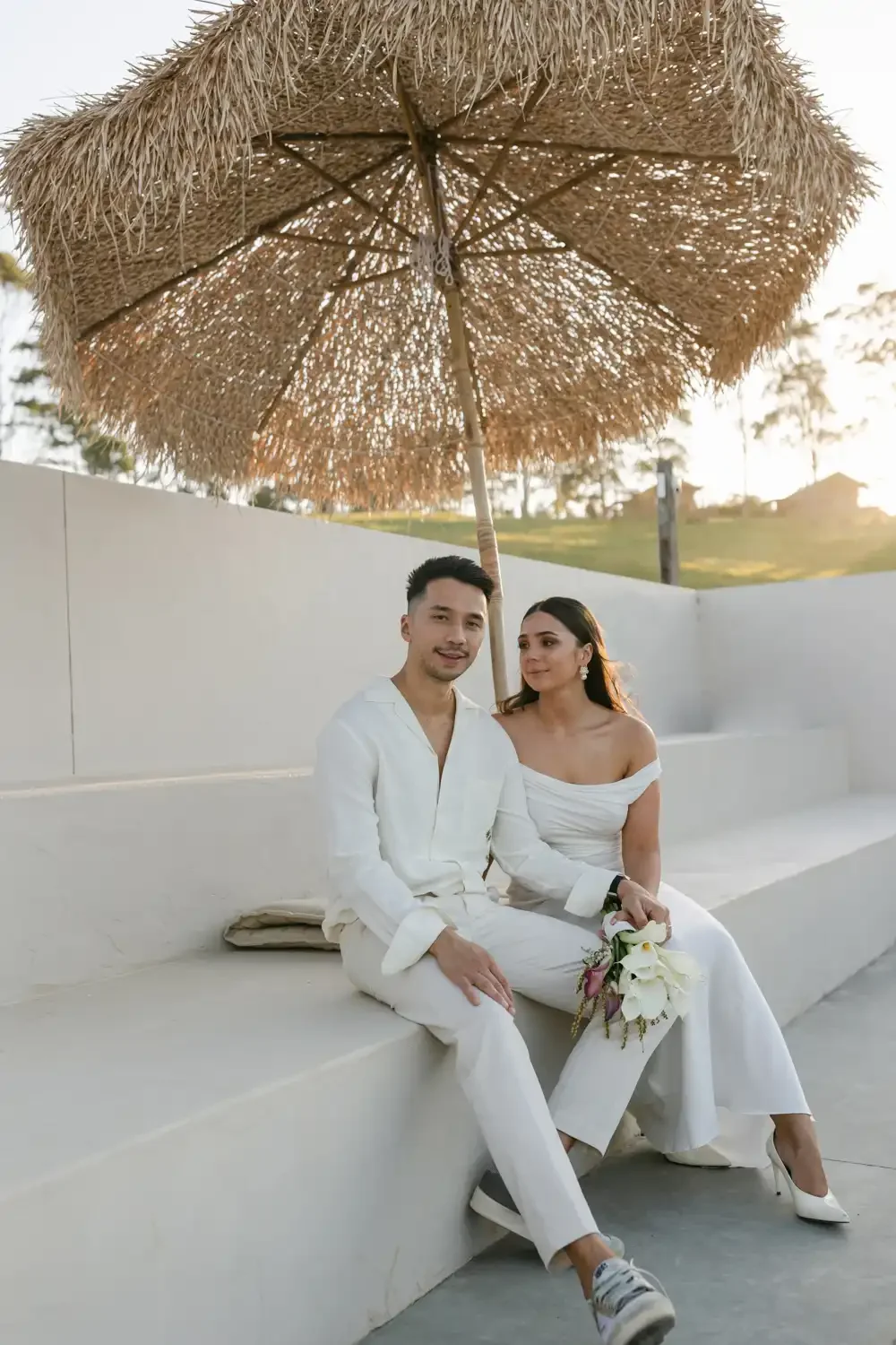 A wedding couple sitting on a white bench under a large straw umbrella outdoors during sunset. The groom is wearing a white shirt and pants, and the bride is dressed in an off-shoulder white gown, holding a bouquet of white flowers.