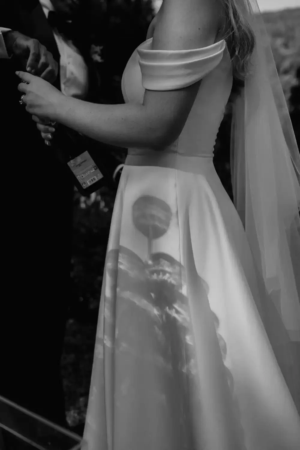 Close-up of a woman wearing an off-shoulder dress with a printed skirt, standing outdoors with trees in the background, holding a phone or small object in her hands.
